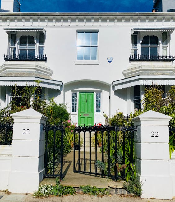 A white Victorian-fronted terraced house with intricate black wrought iron balcony railings and decorative cornices, situated behind a small garden enclosed by a black metal gate. The garden features 