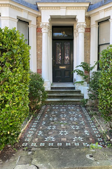A white Victorian-fronted terraced house with intricate black wrought iron balcony railings and decorative cornices, situated behind a small garden enclosed by a black metal gate. The garden features 